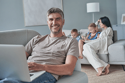 A man smiling at the camera while working on a laptop with his family in the background.